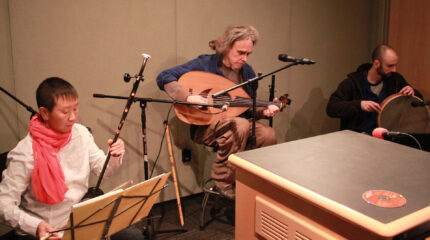 An adult playing an oriental string instrument, an adult playing an ethnic string instrument, and an adult playing a percussion instrument in a radio studio