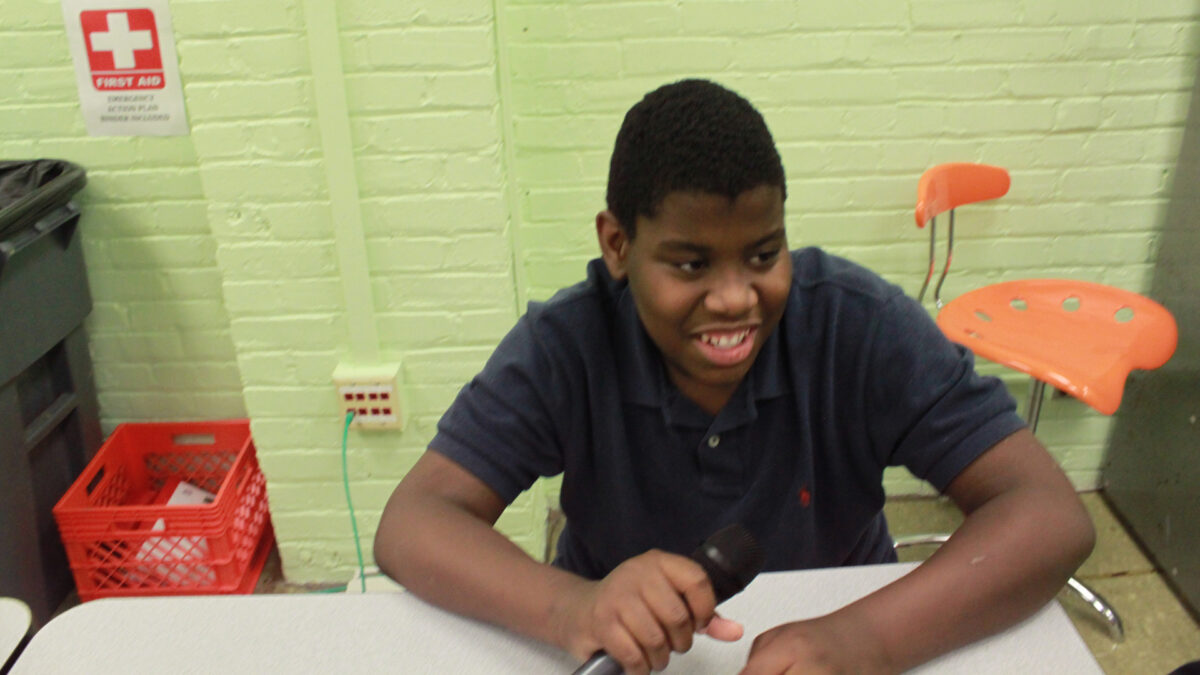 A youth smiling while holding a microphone and sitting at a white desk in a classroom