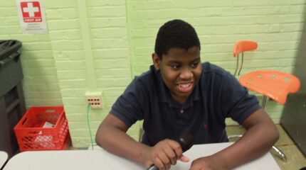 A youth smiling while holding a microphone and sitting at a white desk in a classroom