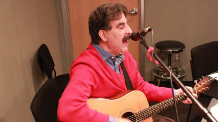 An adult playing acoustic guitar and singing into a microphone in a radio studio
