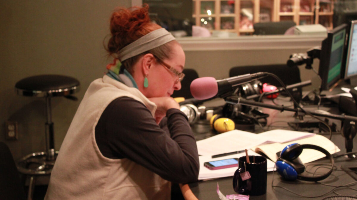 An adult sitting behind a microphone looking down at a smartphone on pieces of paper in a radio studio
