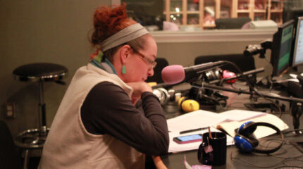 An adult sitting behind a microphone looking down at a smartphone on pieces of paper in a radio studio