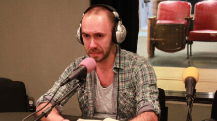 An adult wearing headphones sitting behind a microphone in a radio studio