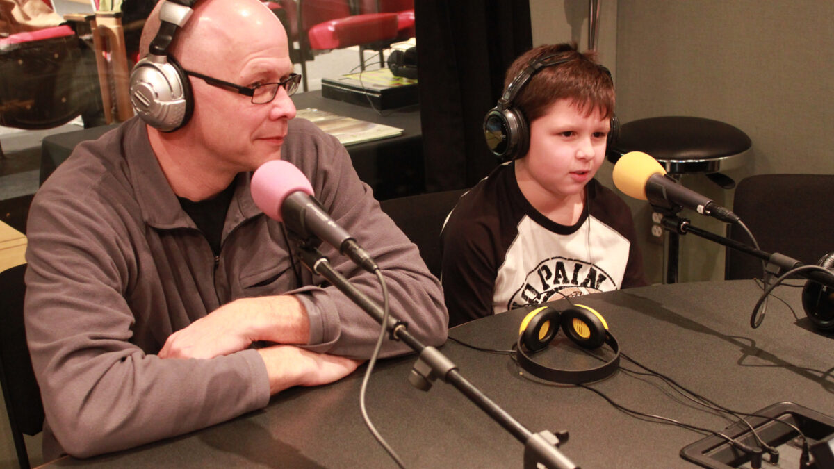 An adult and a youth wearing headphones speaking into microphones in a radio studio