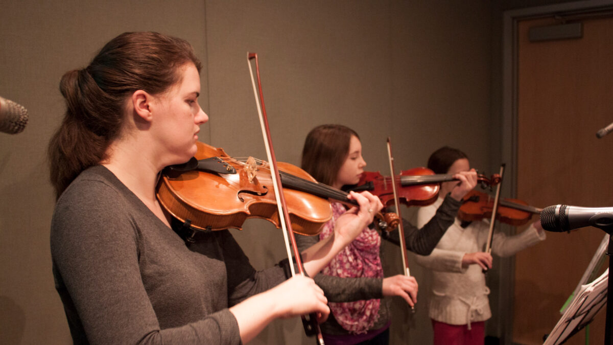 An adult and two youth playing violins and looking at sheet music on music stands
