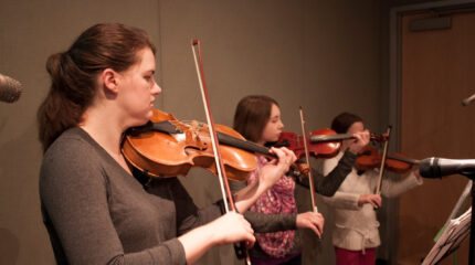 An adult and two youth playing violins and looking at sheet music on music stands