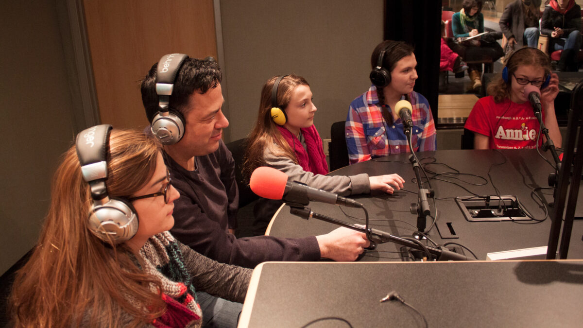 Two adults and three youth wearing headphones speaking into microphones in a radio studio