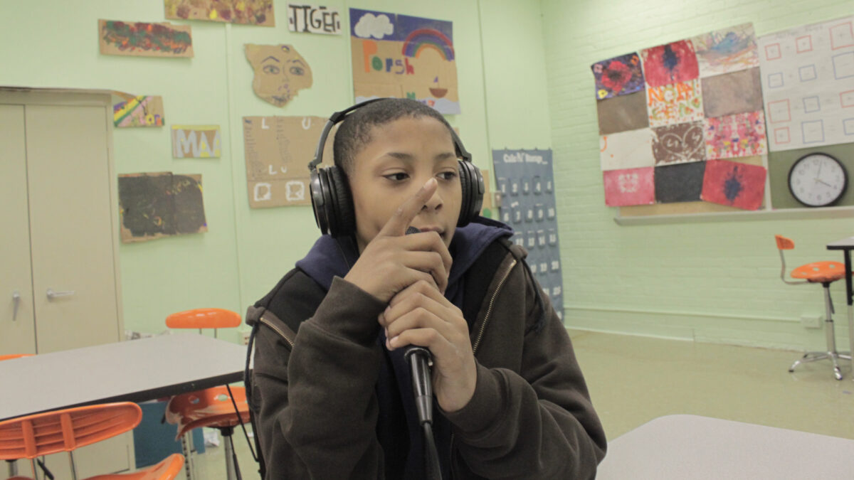 A youth holding a microphone sitting at a desk in a classroom