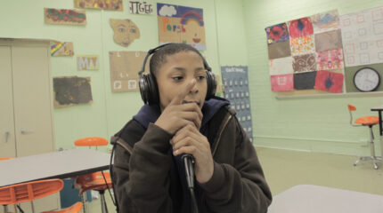 A youth holding a microphone sitting at a desk in a classroom