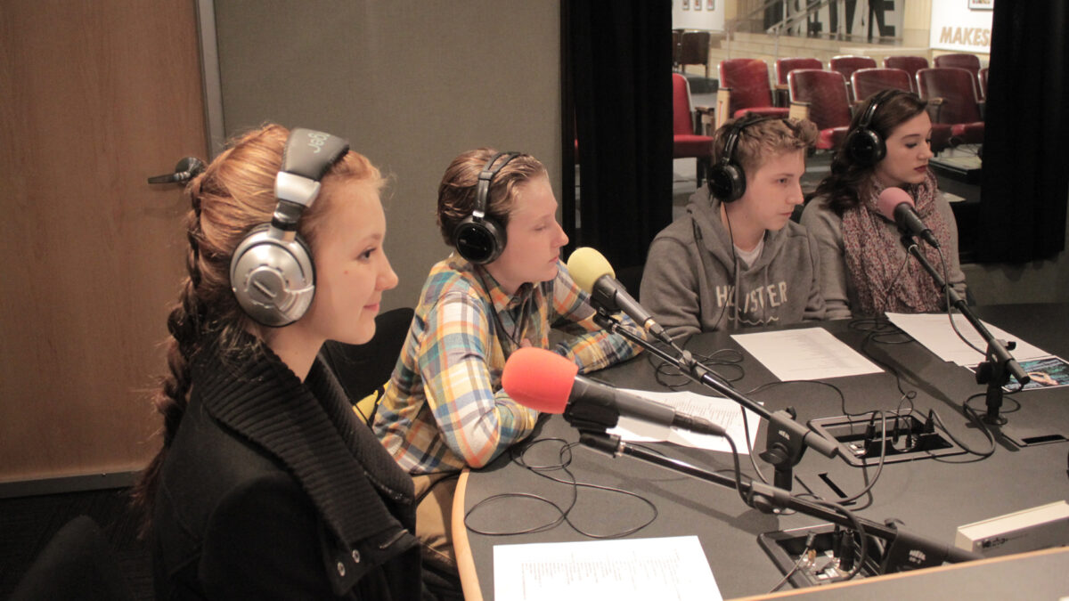 Four teens wearing headphones speaking into microphones sitting behind pieces of paper in a radio studio