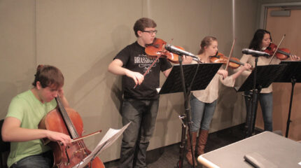 A teen playing cello, a teen playing viola, and two teens playing violins, all looking at sheet music on music stands in a radio studio