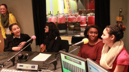 An adult standing in the background watching four teens speaking into microphones in a radio studio