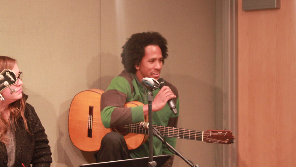 A teen looking at an adult holding an acoustic guitar and speaking into a microphone in a radio studio