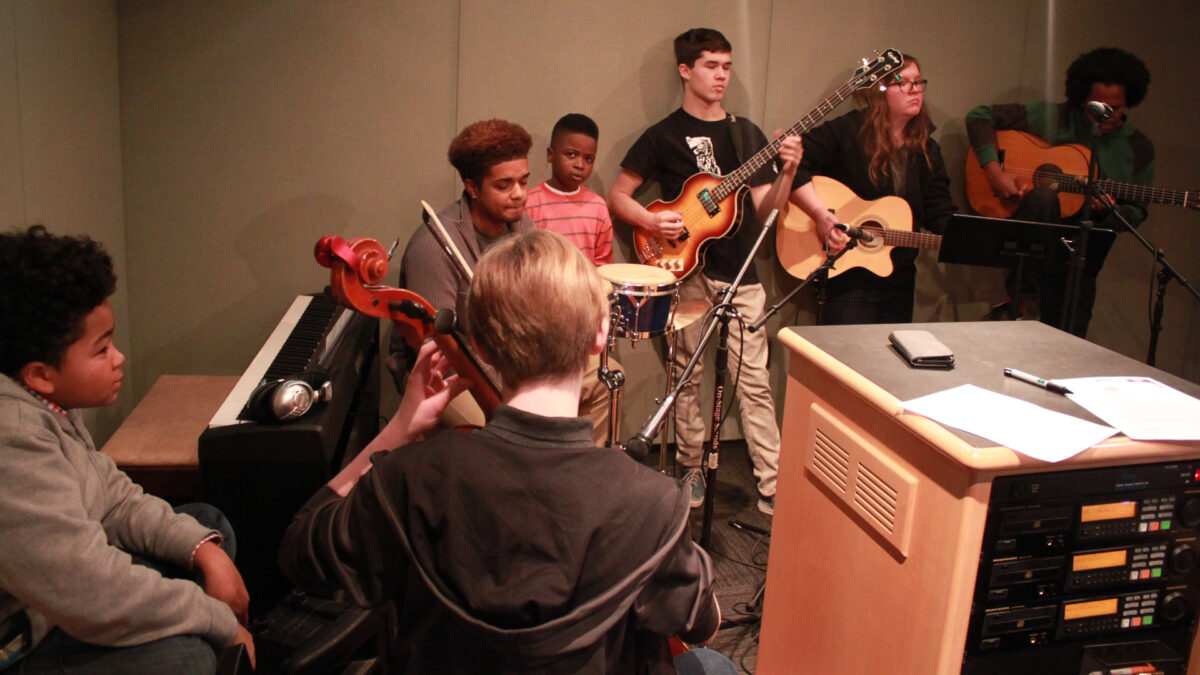 A youth sitting next to a piano keyboard, a teen playing cello, a teen and a youth sitting behind bongos, a teen holding an electric guitar, and a teen and an adult playing acoustic guitars in a radio studio