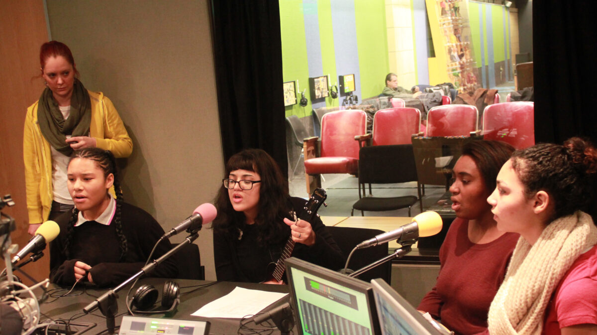 An adult standing in the background watching four teens sing into microphones in a radio studio