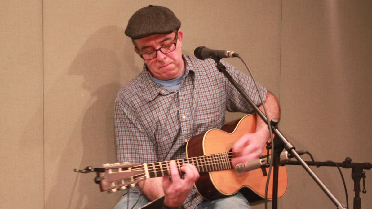 An adult playing acoustic guitar sitting behind a microphone in a radio studio