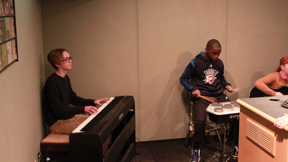 A teen playing a piano keyboard, a teen playing an electric drum set, and a teen playing an acoustic guitar in a radio studio