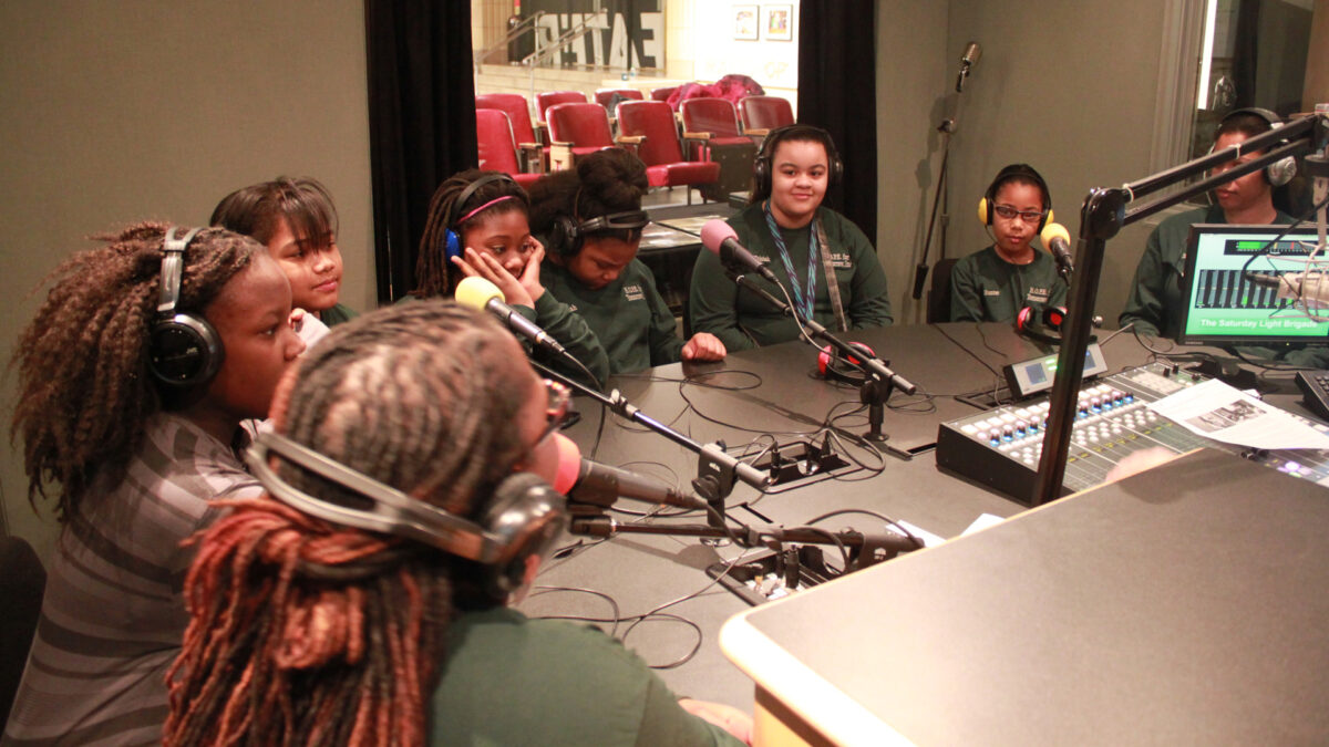 Eight individuals wearing headphones speaking into microphones in a radio studio