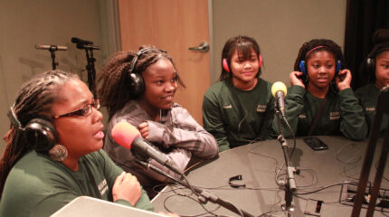 Four youth and an adult wearing headphones and speaking into microphones in a radio studio