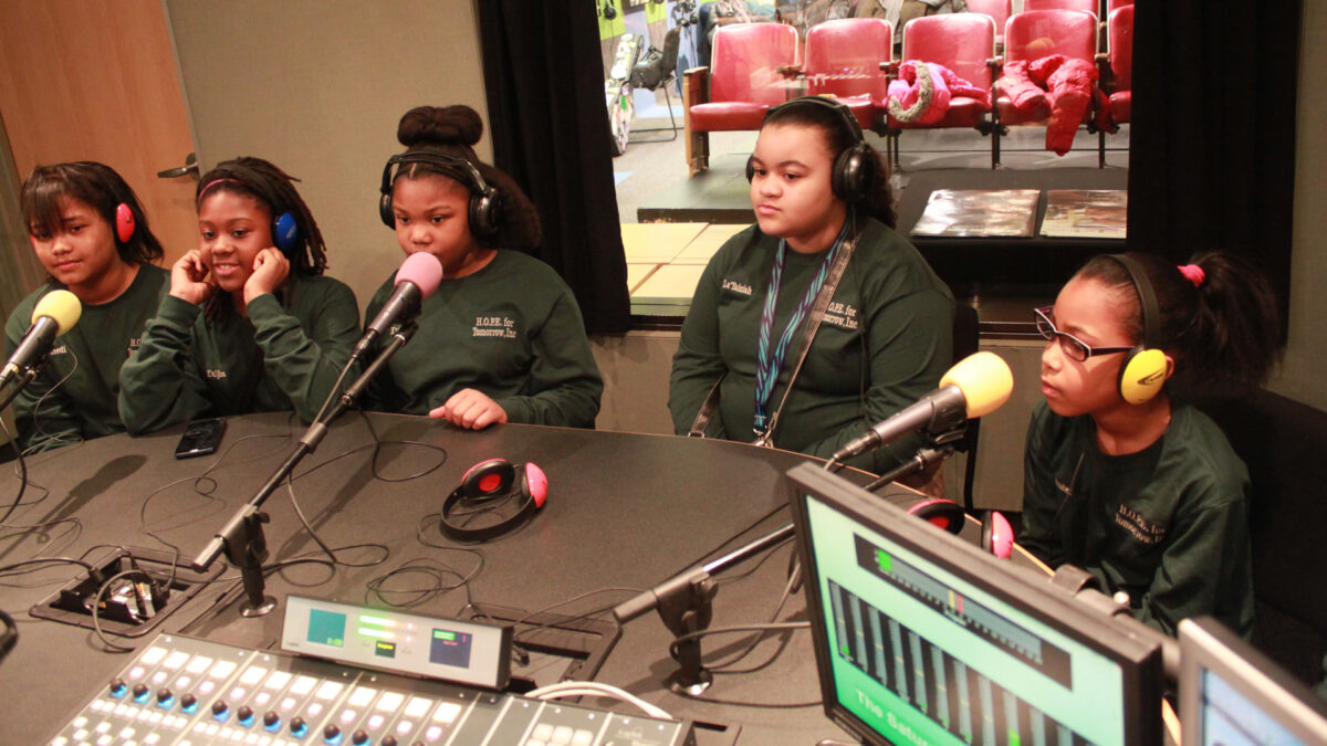 Five youth wearing headphones speaking into microphones in a radio studio