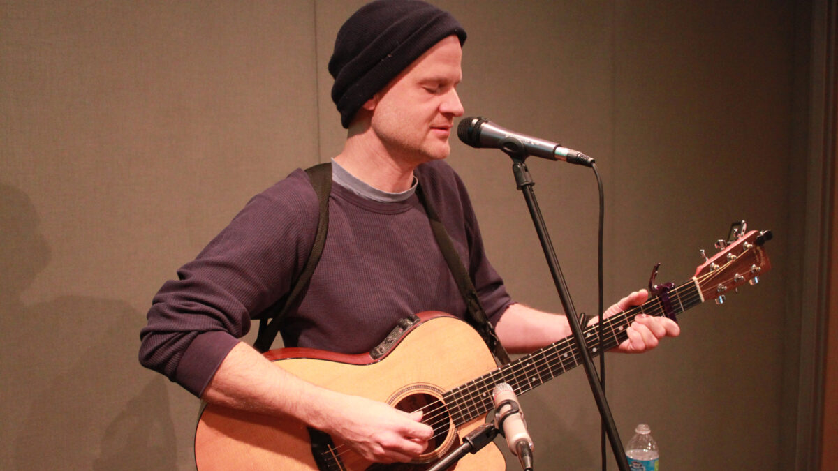 An adult singing into a microphone and playing an acoustic guitar in a radio studio