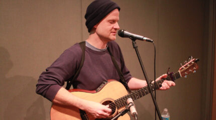 An adult singing into a microphone and playing an acoustic guitar in a radio studio