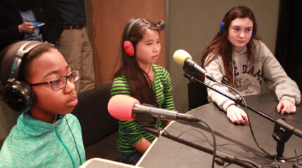 Three youth wearing headphones speaking into microphones in a radio studio