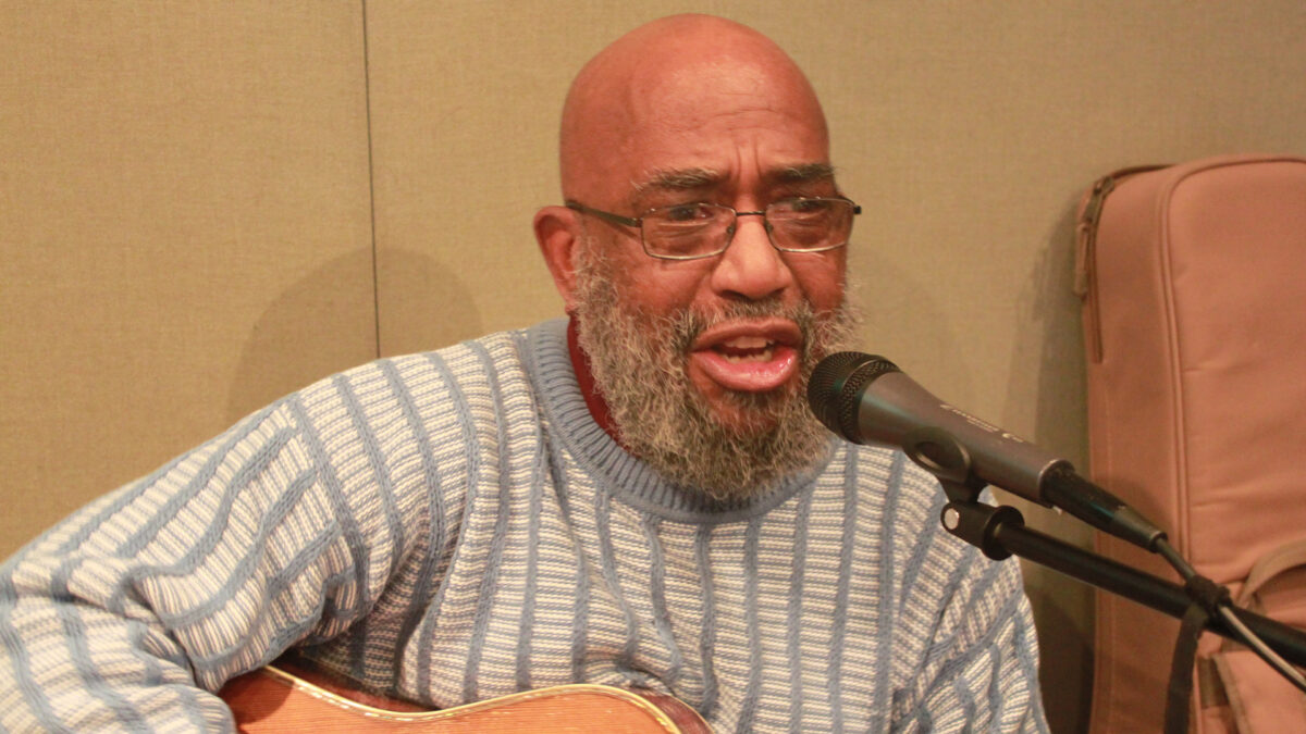 An adult singing into a microphone and holding an acoustic guitar in a radio studio