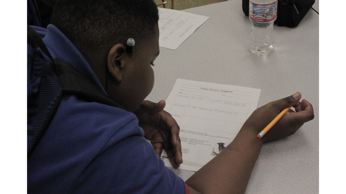 A youth writing on a piece of paper on a gray desk in a classroom