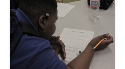 A youth writing on a piece of paper on a gray desk in a classroom