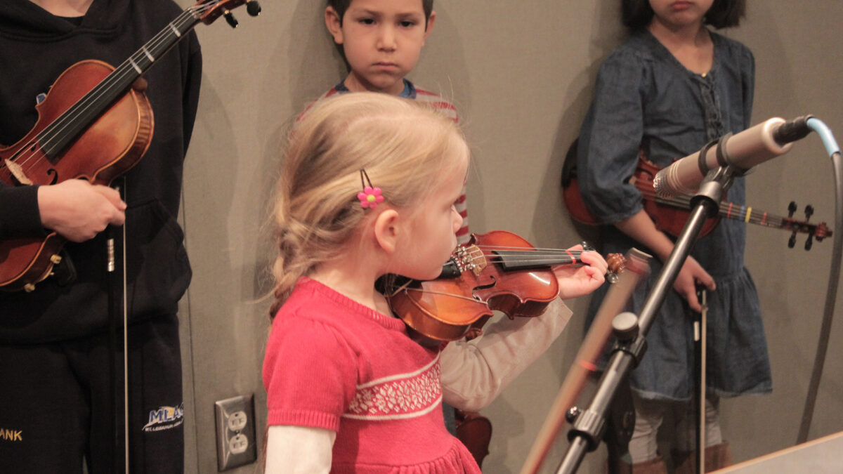 Three youth holding violins watching another youth playing a violin in a radio studio