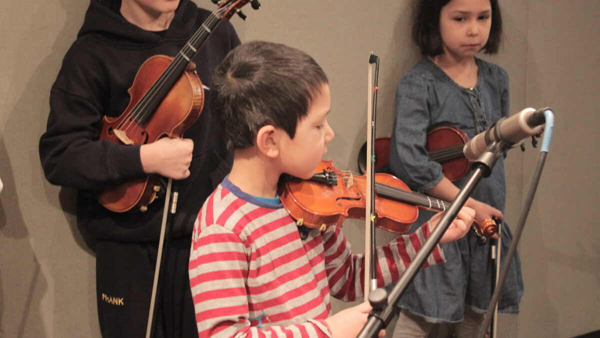 Two youth holding violins watching another youth playing a violin in a radio studio