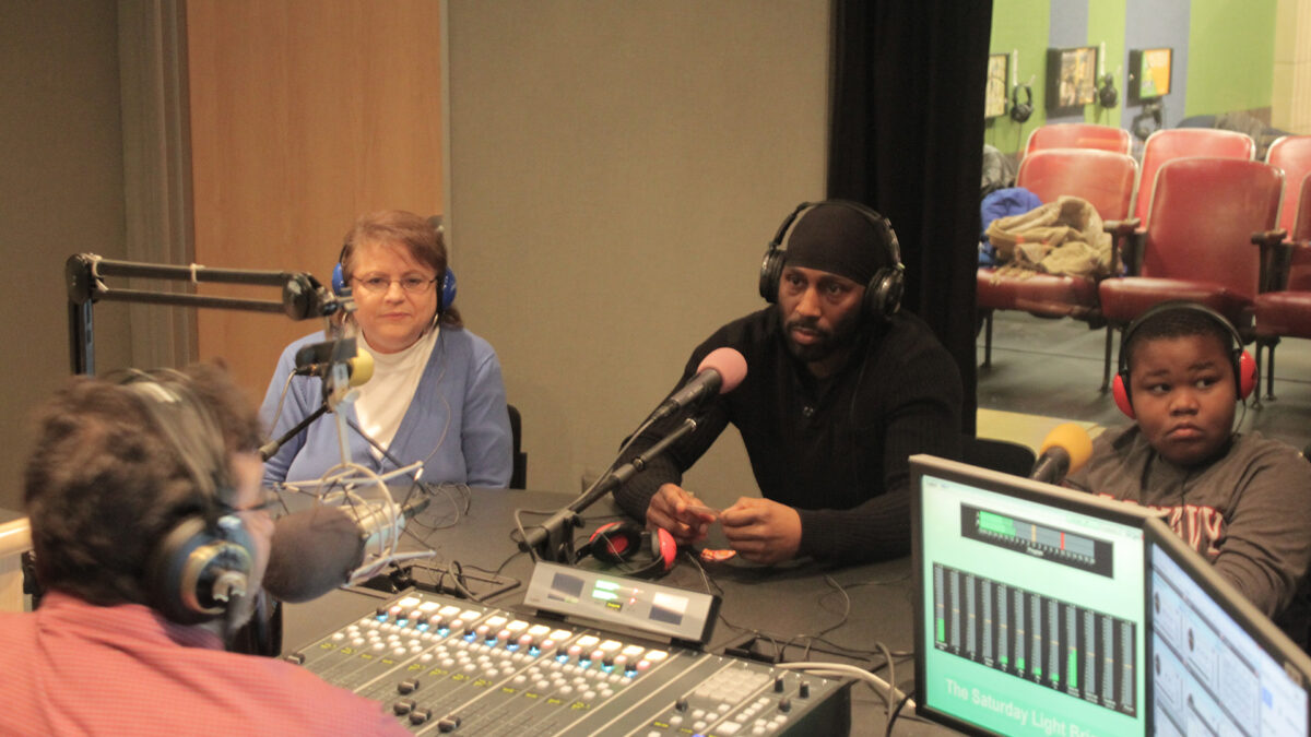 Two adults and a youth wearing headphones sitting behind microphones being interviewed by an adult wearing headphones sitting behind a microphone and a mixer in a radio studio