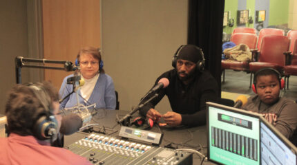 Two adults and a youth wearing headphones sitting behind microphones being interviewed by an adult wearing headphones sitting behind a microphone and a mixer in a radio studio
