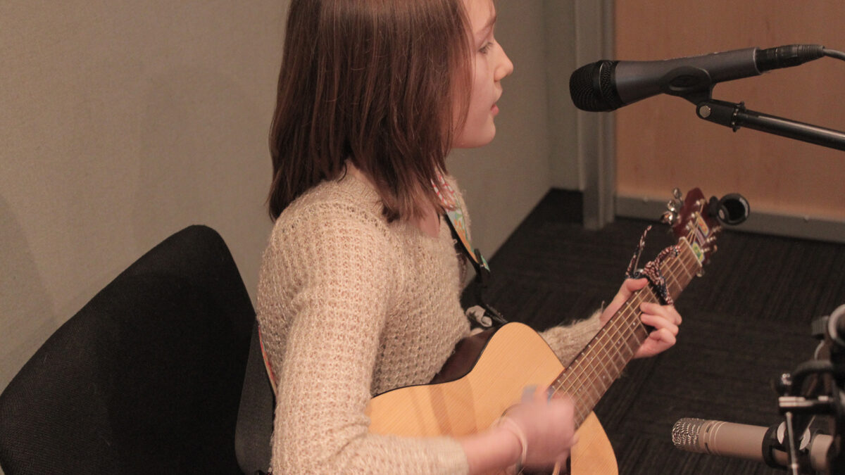 A teen playing an acoustic guitar and singing into a microphone in a radio studio