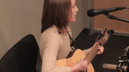A teen playing an acoustic guitar and singing into a microphone in a radio studio