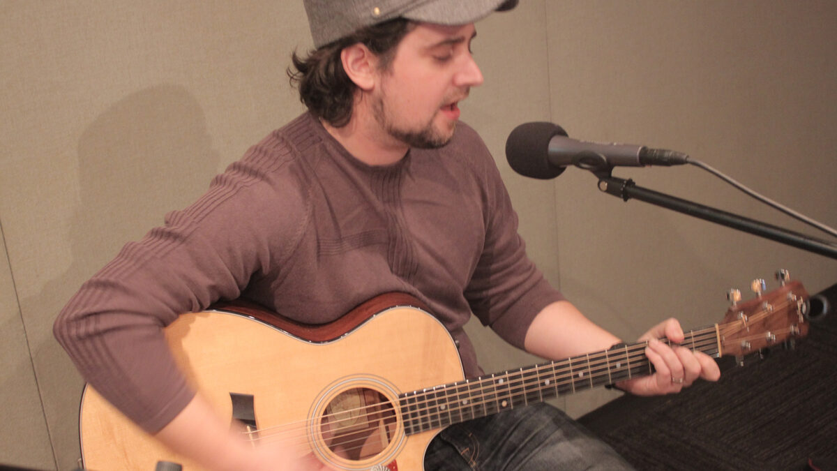 An adult playing an acoustic guitar and singing into a microphone in a radio studio