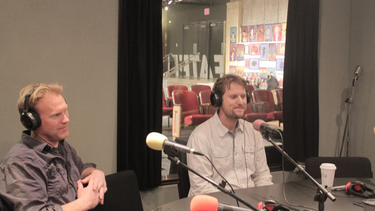 Two adults wearing headphones sitting behind microphones in a radio studio