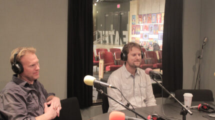 Two adults wearing headphones sitting behind microphones in a radio studio