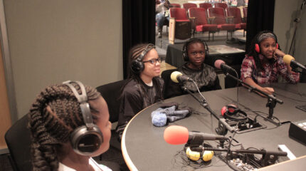 Four youth wearing headphones speaking into microphones in a radio studio