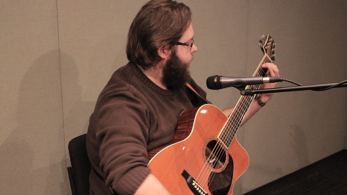 An adult sitting behind a microphone playing an acoustic guitar in a radio studio