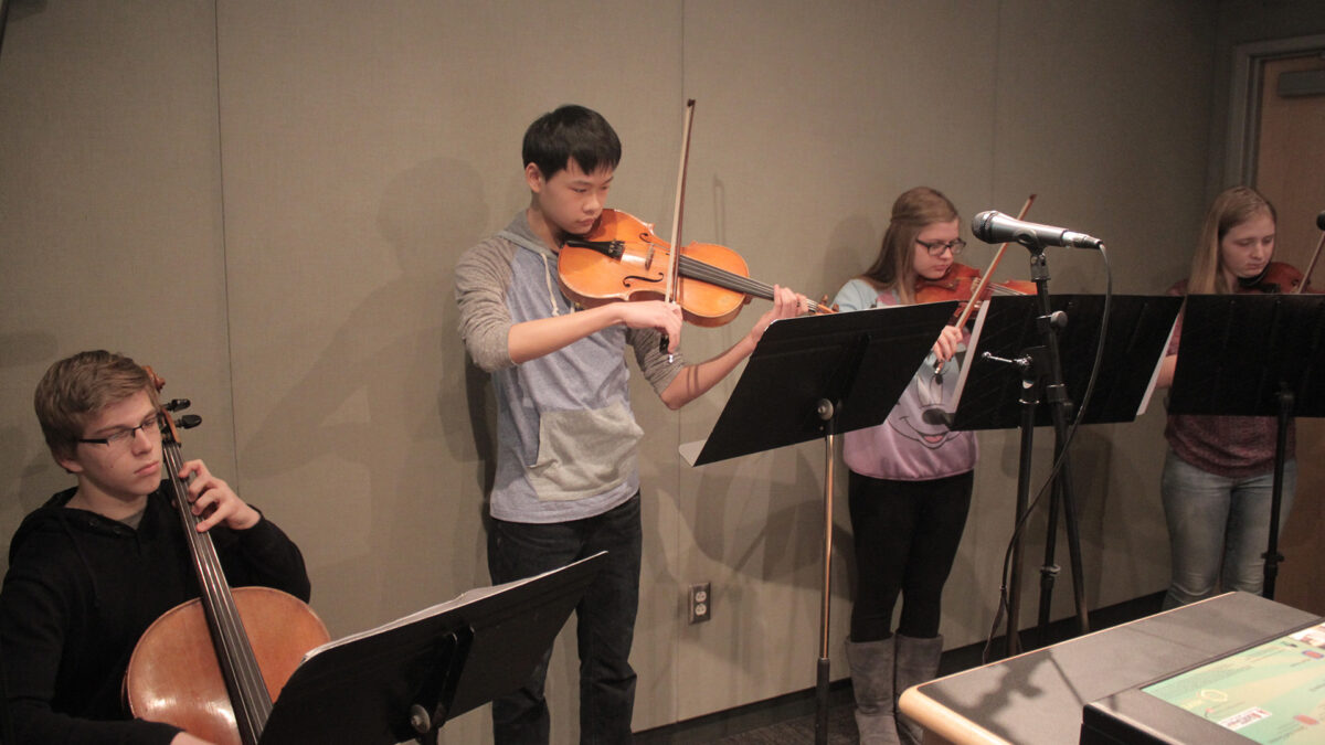 A teen playing cello, a teen playing viola, and two teens playing violins, all looking at sheet music on music stands in a radio studio