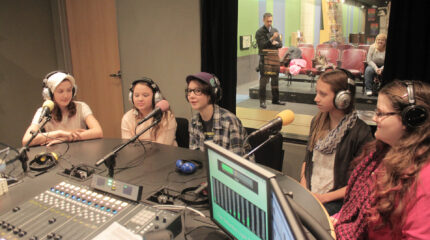 Five youth wearing headphones speaking into microphones being interviewed in a radio studio