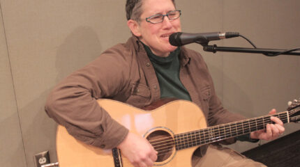 An adult playing acoustic guitar and singing into a microphone in a radio studio