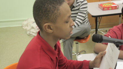 A youth reading a piece of paper and speaking into a microphone in a classroom