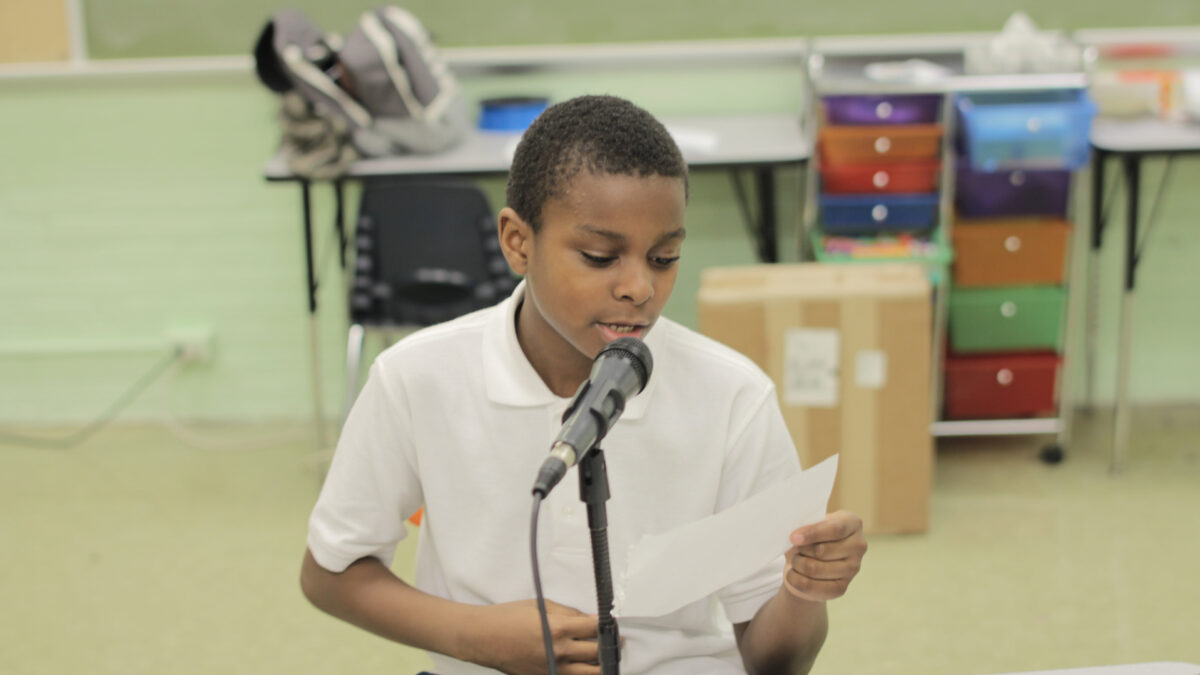 A youth holding a piece of paper and speaking into a microphone in a classroom