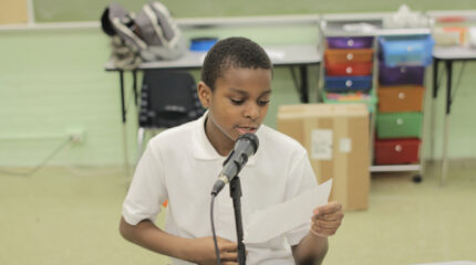 A youth holding a piece of paper and speaking into a microphone in a classroom