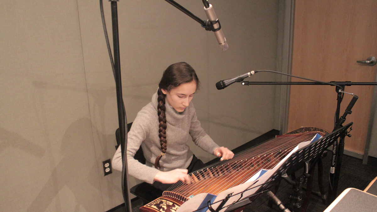 A teen playing a traditional string instrument sitting behind microphones and a music stand in a radio studio