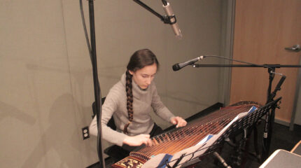 A teen playing a traditional string instrument sitting behind microphones and a music stand in a radio studio