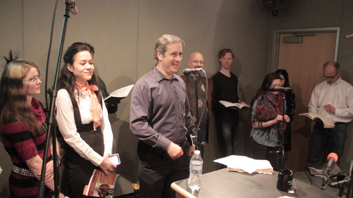 Nine adults standing in front of microphones and looking and books in a radio studio